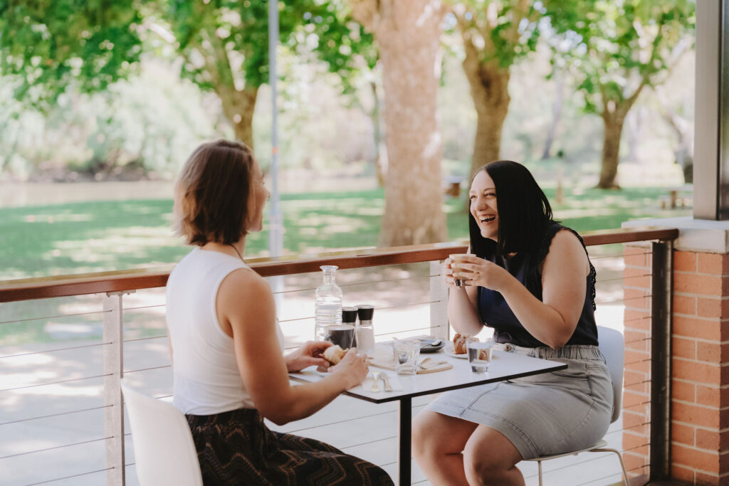 Two women sitting at a table outside. One woman has her back to the camera and is wearing a white blouse. The other woman is wearing a navy blouse and denim skirt and is holding a coffee cup.