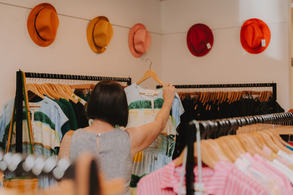 A woman holds up a clue top to look at while browsing clothes in a homeware store. A row of orange and red hued felt hats line the wall