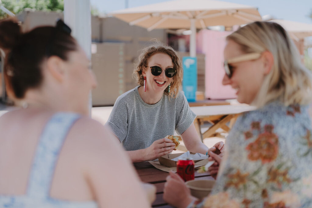 Three women sit at a picnic table under a shade umbrella eating food in carboard take-away containers from nearby vendors.