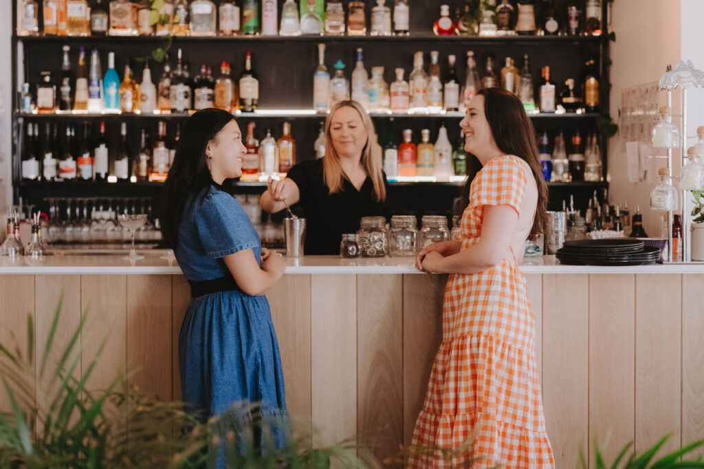 A woman in a blue dress and a woman in a orange check dress are leaning on a bar while a female bartender pours them a drink.