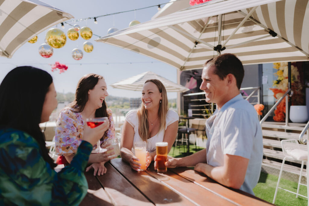 A group of four friends sitting at a wooden table on a rooftop bar under striped umbrellas, enjoying drinks and laughing together on a sunny day. The background shows a cityscape and decorative hanging ornaments above.