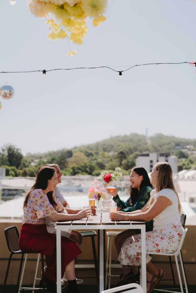 Four friends dressed in bright summery clothing sit at a table at a rooftop bar with drinks in hand. The Monument Hill War Memorial in Albury can be seen atop the hill in the background.