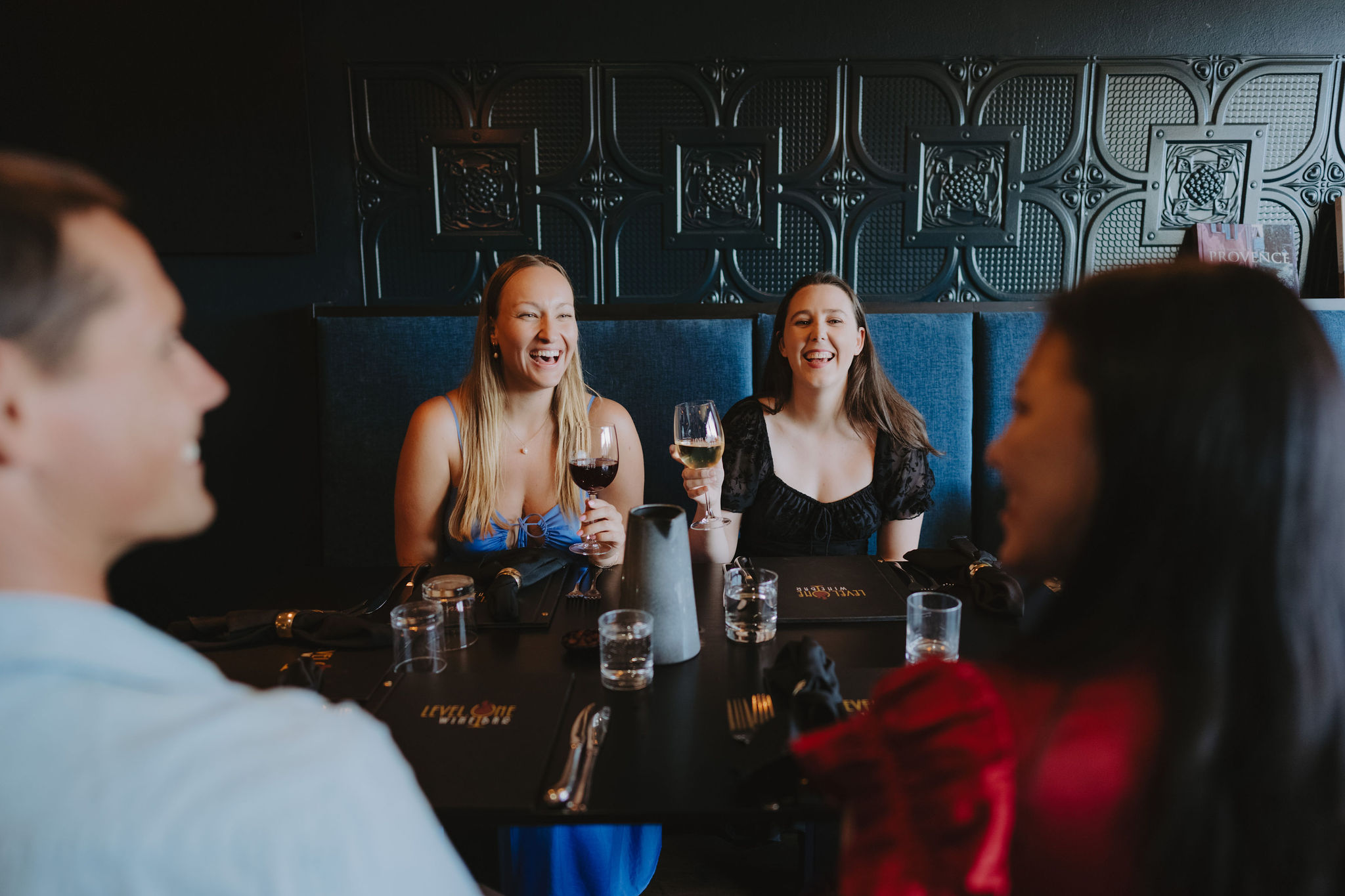 Two women, one with blonde hair and one with brown, laugh with joy as they hold glasses of wine and talk with friends