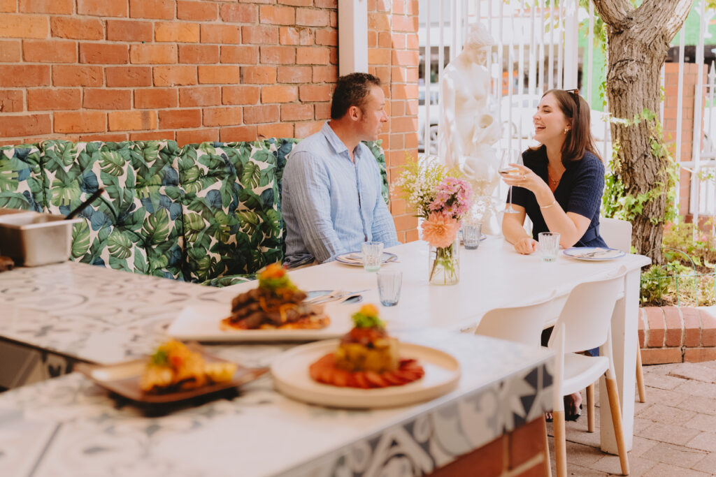 A couple laughing at a table in background with food on a bench in the foreground