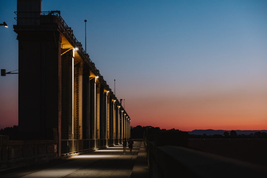 The Hume Dam wall lit with evening lights, with two people walking along it at sunset and a colorful twilight sky in the background.