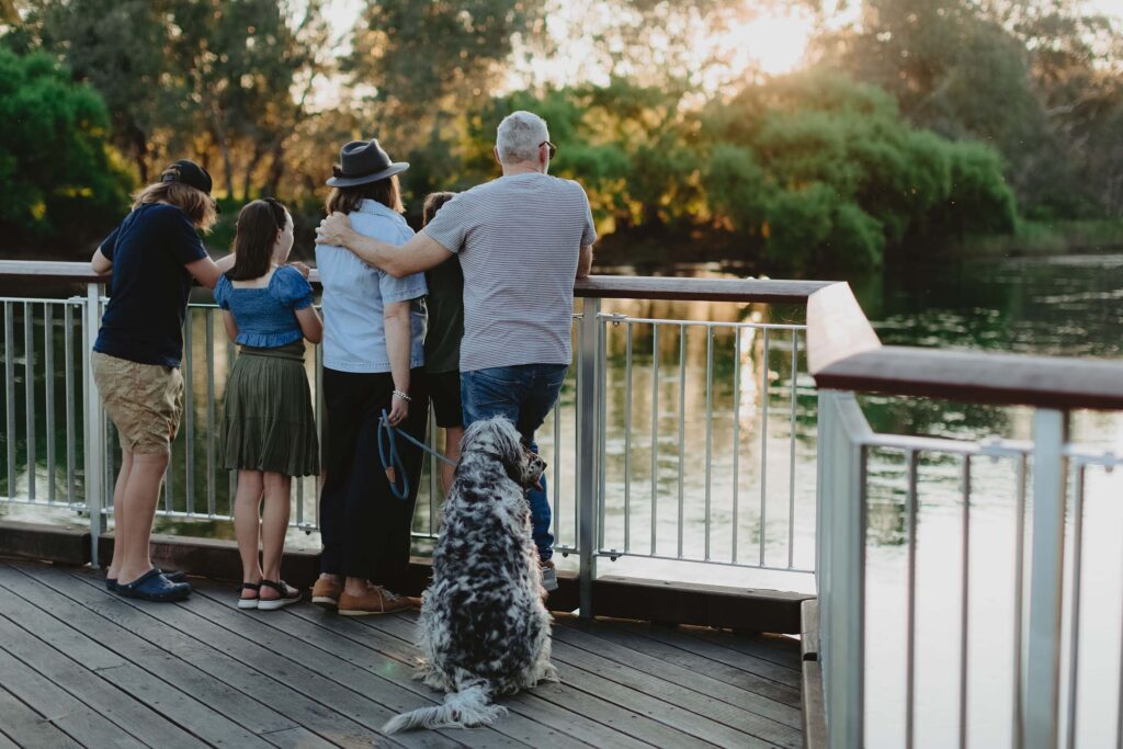 A family of five, including three children and two adults, are standing on a wooden deck by a body of water, looking out at the serene scene. The adults are standing close together, with one arm around the other. A large black-and-white dog is sitting on the deck, facing the water, on a leash held by one of the adults. The setting appears to be during sunset or late afternoon, with warm sunlight filtering through the trees in the background.