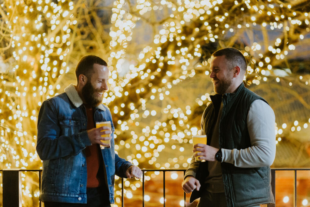 Two men drinking while leaning on a rail in front of a tree covered in fairy lights.