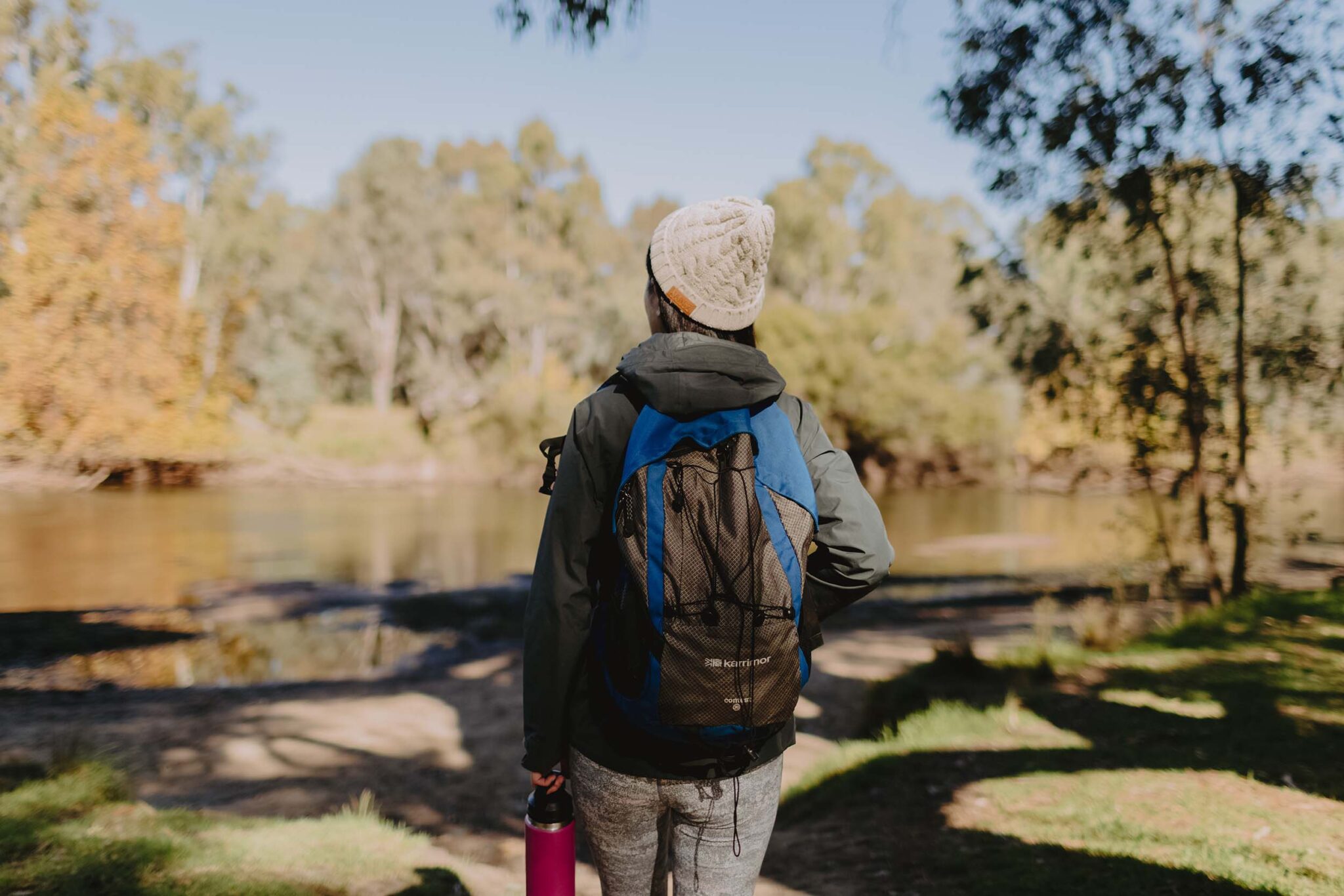 A woman wearing a beanie and a backpack and carrying a pink water bottle stands alone on a walking track looking at the river