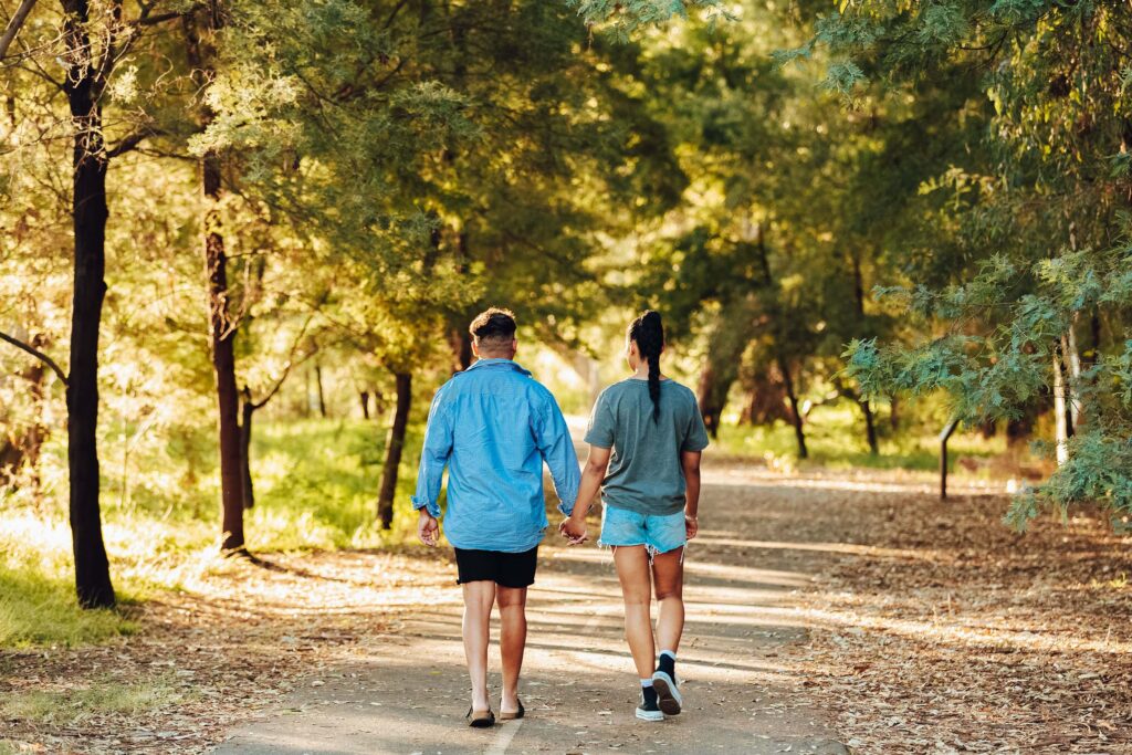 A young couple walk hand-in-hand along a shaded path between acacias and eucalypt trees