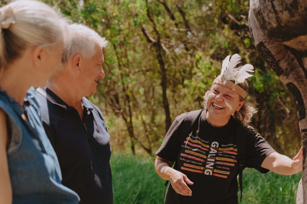 A cheerful person with painted white markings on their face and arms, wearing a black T-shirt with colorful text and a headband adorned with white feathers, is engaging with two older adults outdoors. The older adults, a man and a woman, are listening attentively and smiling. The setting is a sunny, green, wooded area, suggesting a nature walk or cultural tour.