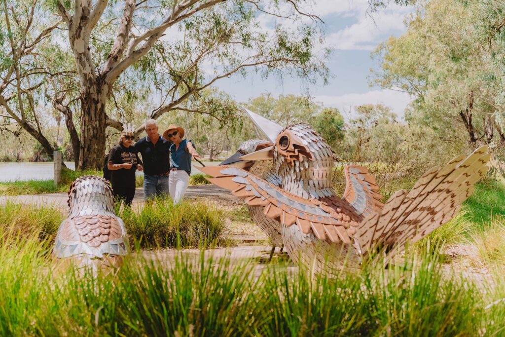 Group of people admiring large metal bird sculptures along the Yindyamarra Sculpture Walk, surrounded by trees and grassy wetlands near a river.