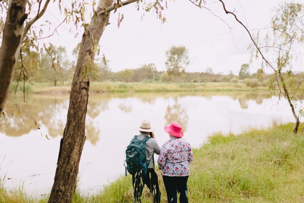 Two people stand side by side at the edge of a calm river surrounded by green grass and trees. One person wears a pink hat and a floral shirt, while the other wears a beige hat, a light gray shirt, and a teal backpack. The scene is peaceful and natural, with soft light filtering through the branches of nearby trees.