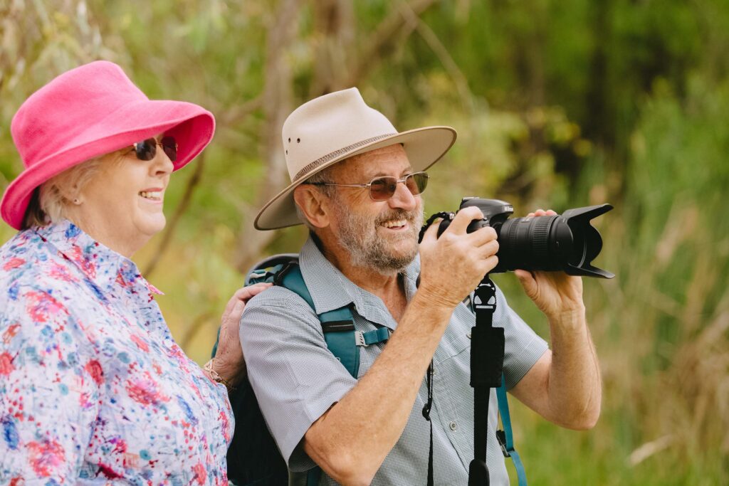 An older couple smiles while birdwatching or taking photos in a natural bush setting; the man holds a DSLR camera.
