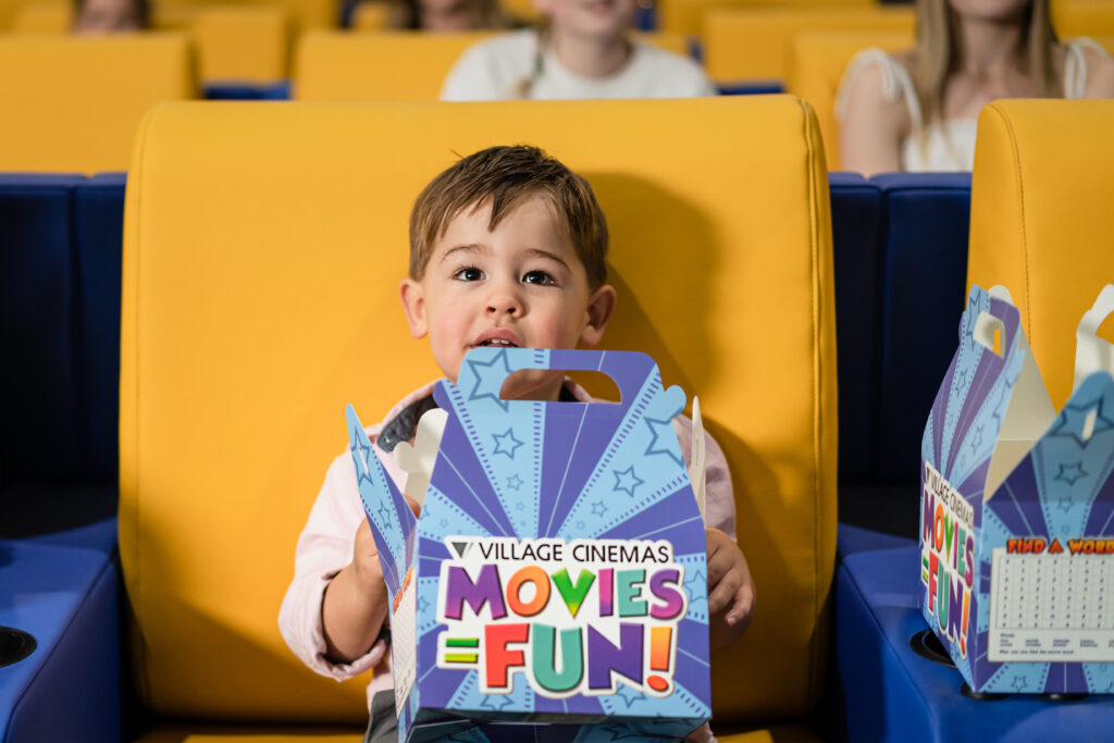A small boy sits in a yellow cinema seat with a carboard meal box on his lap.