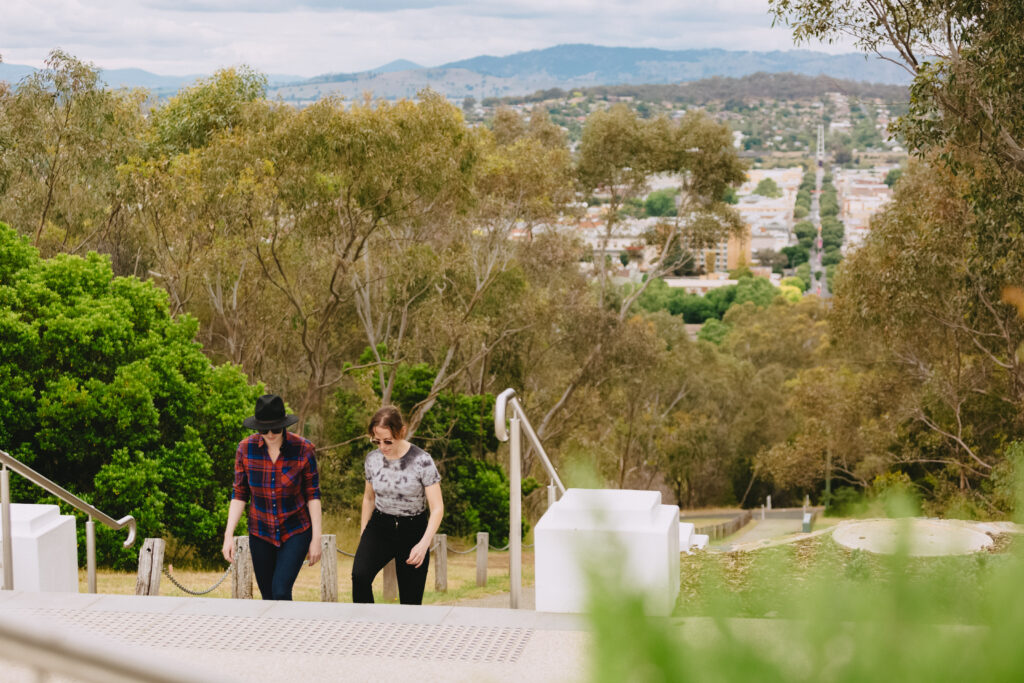 Two women walk up a set of outdoor stairs surrounded by greenery, with a scenic view of a town and distant hills in the background.