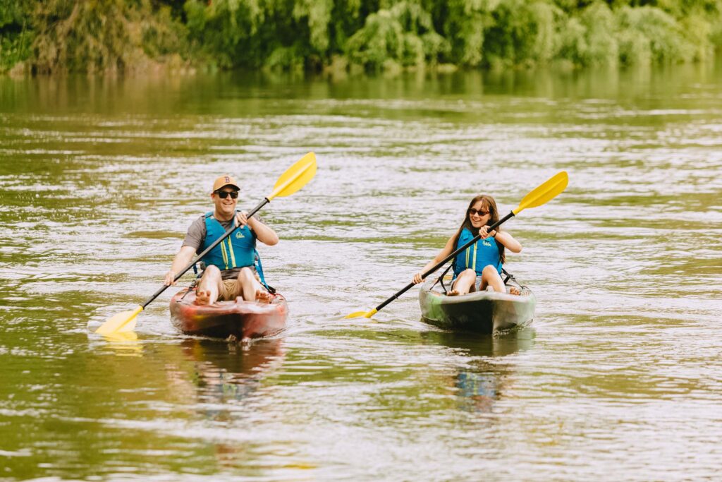Two people kayaking on a calm river, both wearing life jackets and sunglasses, smiling as they paddle side by side with bright yellow paddles.