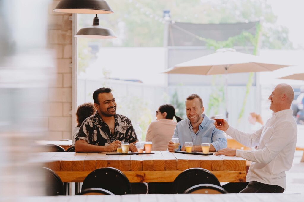 Three men sit around a wooden table, smiling and enjoying a beer tasting at a brewery. The man on the left wears a black floral shirt, the man in the middle wears a light blue shirt, and the man on the right, raising a glass, wears a white shirt. The setting is bright and casual, with large umbrellas and other patrons in the background.