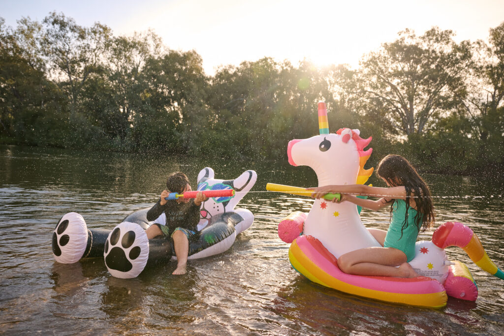 A young boy dressed in black swim wear is sitting on a white and blue bear floaty while a young girl in green swimmers is sitting on a unicorn floaty. Both are spraying each other with water sprays.
