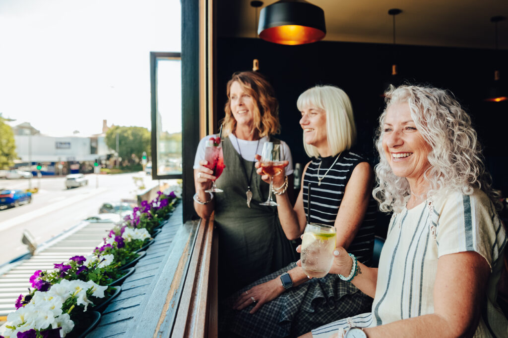 Three women sitting by an open window at a bar, smiling and holding drinks while looking out toward the street. Flower boxes with purple and white blooms line the window ledge, and the bright daytime street scene is visible outside.