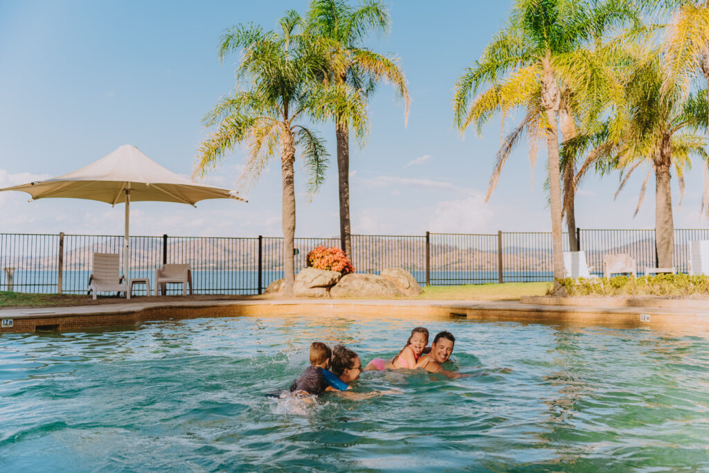 A family of four is swimming in a blue pool. The young children are riding on the backs of their parents.