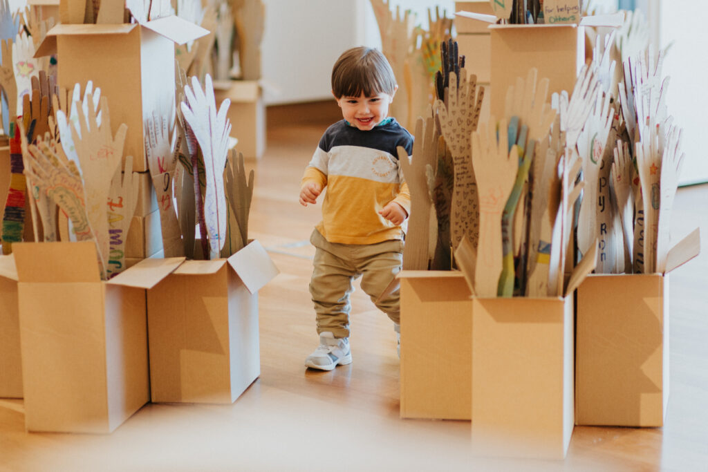 A toddler in a grey, white and yellow stripped shirt is playing between some boxes with a smile on his face