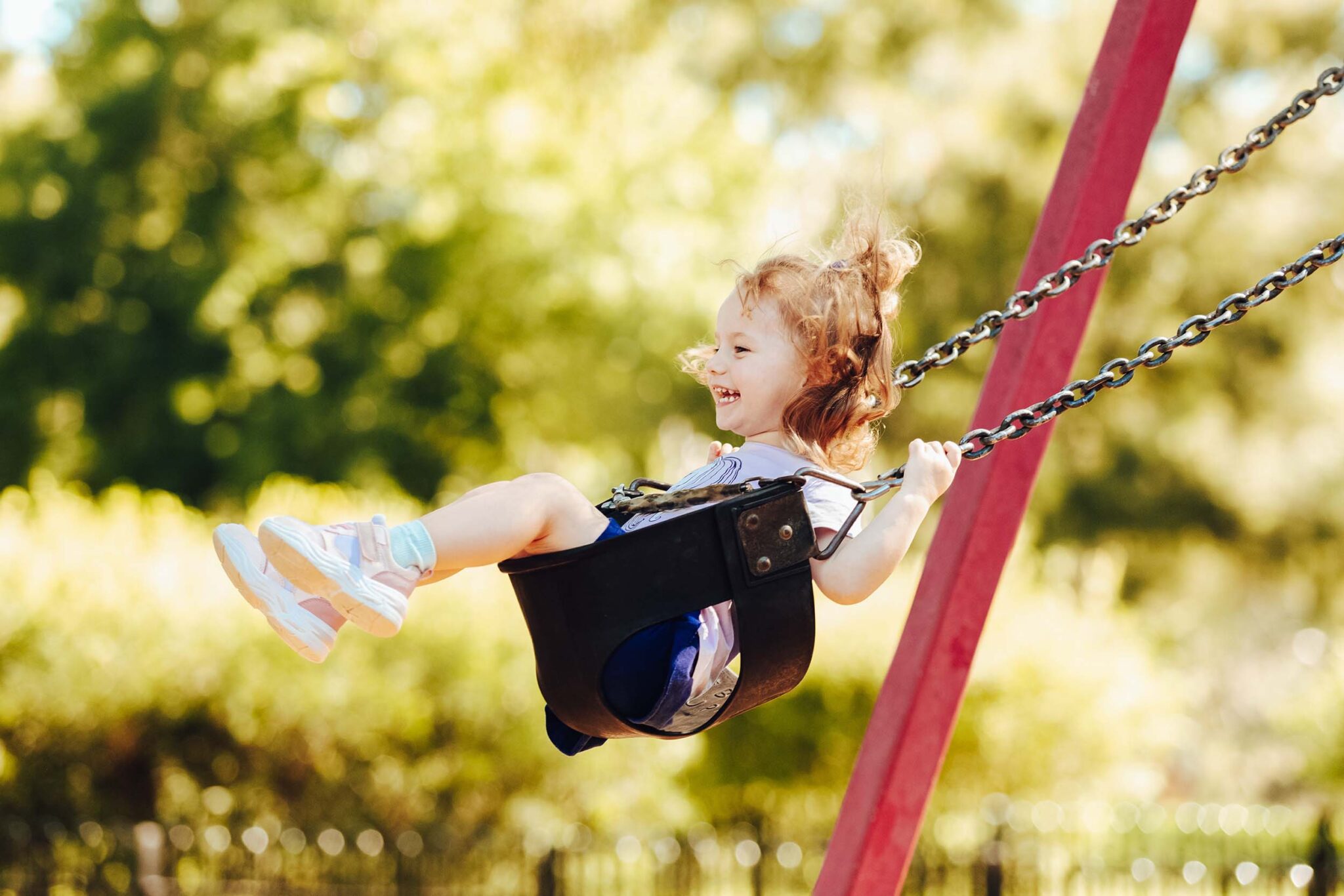 Young girl with a big smile buckled into toddler swing, captured at the top of the swing's arc
