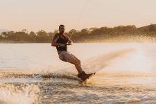 A man wearing a life jacket and grey boardshorts rides a wakeboard on Lake Hume at dusk.