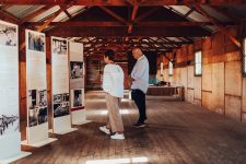 An older couple read a display about migrants in an old sleeping hut with timber floors and basic camp beds.