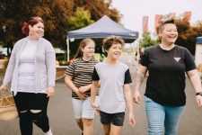 Two women and two children smile as they walk through a market. There is a marquee behind them. One of the women has red hair and carries a woven basket.