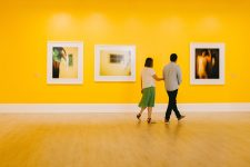 A woman in a green skirt and a man in a blue shirt with black pants walk through an art gallery with bright yellow walls