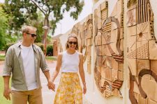 A smiling man and woman walk hand-in-hand along a pathway beside a wall adorned with textured, geometric clay tile art. Both are wearing sunglasses; the man is dressed casually in a white t-shirt, light green button-up shirt, and tan shorts, while the woman wears a white sleeveless top and a yellow floral skirt. Trees and greenery are visible in the background, suggesting a warm, sunny day.