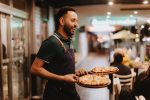 A smiling man wearing an apron carries two pizzas on wooden serving trays across a footpath to outdoor diners at night.