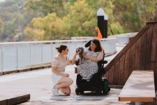 A woman in a motorised wheelchair is stopped on a boardwalk and she holds a small grey dog. A woman kneels beside her to pat the dog. There are eucalypt trees in the background.