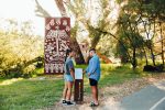 Two people stand in front of an Aboriginal art installation and interpretive sign along a tree-lined riverside path, engaging with Indigenous culture and history in a natural setting.